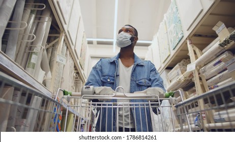 African man wearing protection facemask choosing wallpaper in hardware store. Low angle view of afro guy pushing shopping cart buying materials in house improvement store wear safety mask and gloves - Shutterstock ID 1786814261