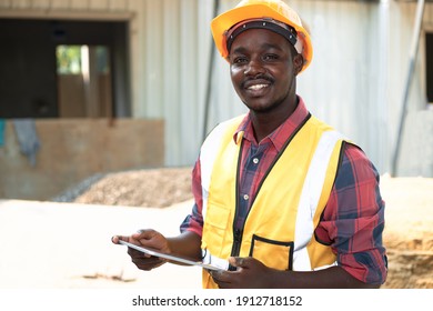An African Man Wearing A Yellow Vest And A Hard Hat Holding A Tablet At A Construction Site.