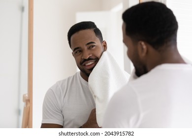 African man in white t-shirt stand in bathroom look in mirror wipes his face with towel after shave, skin care facial procedure smiles feels satisfied. Male beauty cosmetics, skincare, hygiene concept - Powered by Shutterstock