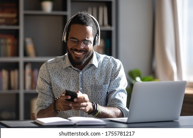 African Man Worker Or Student Sit At Desk Distracted From Study Or Work Hold Smartphone Wear Headphones Hearing Voice Audio Message From Friend, Enjoy Free Time Use Modern Gadget Wireless Tech Concept