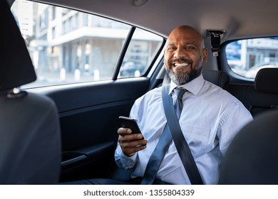 African Mature Businessman Holding Smartphone While Looking At Camera. Successful Entrepreneur Wearing Blue Tie Sitting In Car Using Mobile Phone. Happy Formal Black Business Man Traveling In Taxi.