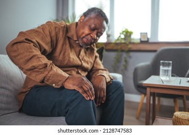 An African mature man suffers from knee pain while sitting on a couch. A mature man massages his aching knee. A man who suffers from knee pain at home, up close. Puts both hands on an aching knee.