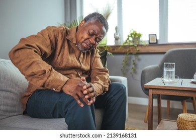 An African mature man suffers from knee pain while sitting on a couch. A mature man massages his aching knee. A man who suffers from knee pain at home, up close. Puts both hands on an aching knee.