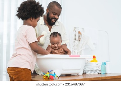 African middle aged father bathing adorable newborn baby daughter in bathtub at home. Child boy help dad cleaning, washing little sister with sponge in bath. Kid hygiene and health care with family - Powered by Shutterstock