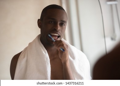 African Millennial Guy After Shower Brushing Teeth Looking In Mirror Standing In Bathroom, 30s Shirtless Ma, Towel On Shoulders, Cleaning Teeth, Morning Routine Oral Hygiene, Healthy Lifestyle Concept