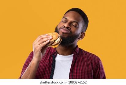 African Millennial Guy Smelling Tasty Burger Standing On Yellow Studio Background. Black Man Enjoying Eating Hamburger. Unhealthy Nutrition Habit, Overeating. Junk Food Lover Concept