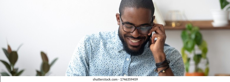 African millennial man holding smartphone enjoy distant conversation seated indoors, horizontal photo banner for website header design. Successful businessman make business call, informal talk concept
