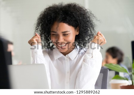 African mixed race female sitting at desk in office looking on laptop screen feels happy. Head shot portrait black millennial employee received long awaited great news about reward or job advancement
