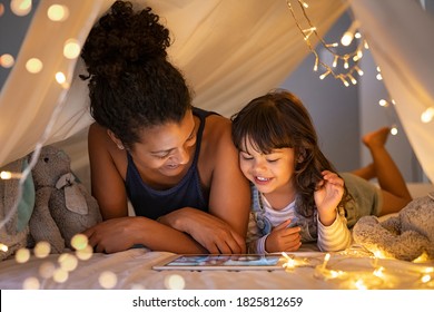 African mother and cute smiling girl using digital tablet while lying in illuminated tent in kid bedroom. Cheerful ethnic woman and lovely daughter on video call under a cozy hut. Lovely little girl.