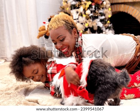 African mother and daughter play and laugh on a woolen carpet with a funny dog in Santa Claus clothes against the backdrop of a Christmas tree and fire in the fireplace