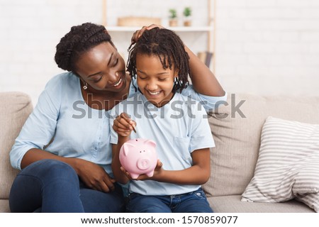 African Mother And Daughter Putting Coins Into Piggy Bank At Home. Free space