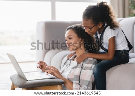 African mother and daughter watching movie together on laptop at home. Mom freelancer trying to concentrate on distant remote work while small daughter disturbing her. Maternity leave concept