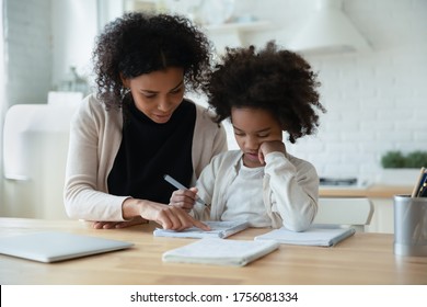 African mother helps with task little schoolgirl daughter do together schoolwork, parent explain subject, focused child listen to mum sit at table at home in kitchen. Education, homeschooling concept