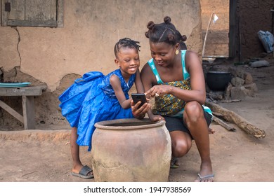 An African mother with her children bending low beside a big clay plot in front of a village mud house with a mobile phone in the mothers hand 