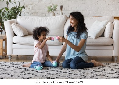 African mother and little daughter holding plastic toy teacups enjoy tea ceremony play together sit on carpet in warm living room. Dishware shop advertisement, weekend playtime with children concept - Powered by Shutterstock