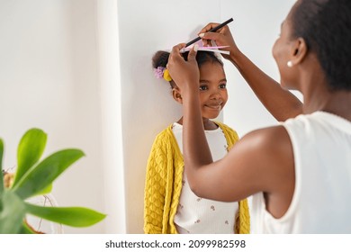 African mother measuring daughter height and marking it on wall at home. Happy black mother measuring cute little girl height at home using book and pen. Cheerful mature woman measuring her daughter. - Shutterstock ID 2099982598