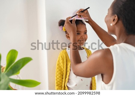 African mother measuring daughter height and marking it on wall at home. Happy black mother measuring cute little girl height at home using book and pen. Cheerful mature woman measuring her daughter.