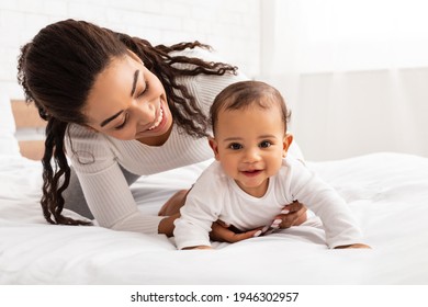 African Mother Playing With Cute Baby Daughter Helping Her Crawl On Bed In Bedroom At Home. Young Mommy Enjoying Motherhood Routine And Child Care. Maternity Leave Lifestyle