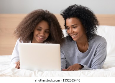 African Mother And Teen Daughter Spending Lazy Weekend Together Lying On Bed At Home Or Hotel Room Use Computer Browse Web Watching Video Interesting Vlog Search Best Offers Via Internet Buying Online