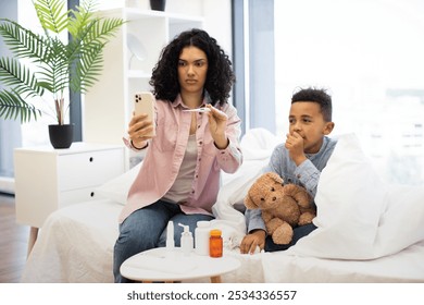 African mother uses smartphone for video consultation with doctor for sick young son on bed. Child holds teddy bear, covered with blanket, while mother checks temperature with thermometer. - Powered by Shutterstock