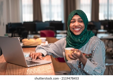 African Muslim Business Woman Wearing A Green Hijab Drinking Tea While Working On Laptop Computer In Relaxation Area At Modern Open Plan Startup Office.