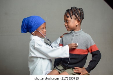 African Nigerian Girl Child With Nose Mask And Stethoscope, Playing Medical Expert Like Doctor And Nurse With Kid Patient Sitting In Front Of Her