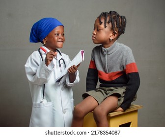 African Nigerian Girl Child With Nose Mask And Stethoscope, Playing Medical Expert Like Doctor And Nurse With Kid Patient Sitting In Front Of Her