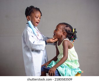 African Nigerian Girl Child With Nose Mask And Stethoscope, Playing Medical Expert Like Doctor, Nurse And Dentist With Kid Patient Sitting In Front Of Her