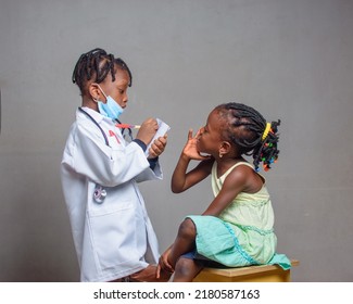 African Nigerian Girl Child With Nose Mask And Stethoscope, Playing Medical Expert Like Doctor And Nurse With Kid Patient Sitting In Front Of Her