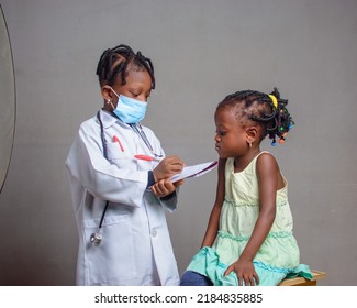 African Nigerian Girl Child With Nose Mask And Stethoscope, Playing Medical Expert Like Doctor And Nurse With Kid Patient Sitting In Front Of Her