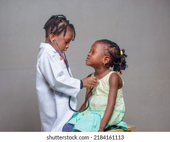 African Nigerian Girl Child With Stethoscope, Playing Medical Expert Like Doctor And Nurse With Kid Patient Sitting In Front Of Her