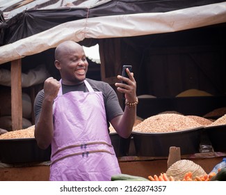 An African Nigerian male trader, seller, business man or shop owner, having an apron on his body and amazed as he stares into the smart phone in his hand in a market