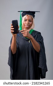 African Nigerian Muslim Lady, Student Or Graduand In Hijab And Graduation Gown, Points To The Smart Phone In Her Hand