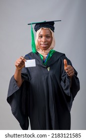 African Nigerian Muslim Lady, Student Or Graduand, Wearing An Hijab Head Scarf And Graduation Gown, Holding A Small White Card And Doing Thumbs Up Gestures With Her Second Hand