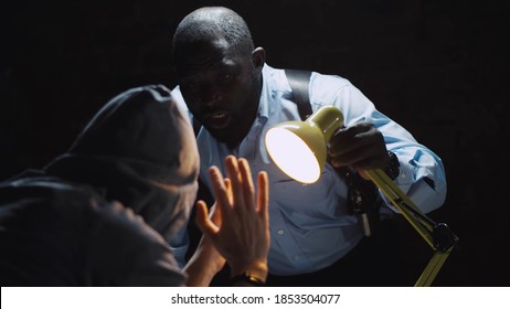 African officer interrogating man suspected of crime glowing light of lamp into face. Afro-american sheriff questioning arrested man in dark room of police department