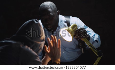 African officer interrogating man suspected of crime glowing light of lamp into face. Afro-american sheriff questioning arrested man in dark room of police department