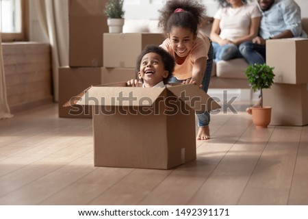 African older sister riding younger cute brother who is sitting in cardboard box, on background big carton boxes stuff and parents sitting on couch enjoy new modern house resting at moving day concept