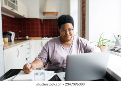 African plus size aged woman sitting in front of laptop planning purchases, making list before going to supermarket, using online application to compare prices, searching for sales, special offers - Powered by Shutterstock