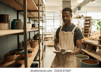 African potter doing an inventory of pots on shelves in a ceramics workshop - Powered by Shutterstock