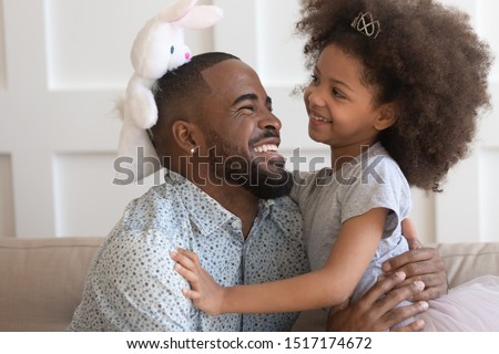 African preschool daughter in crown like princess holding rabbit soft toy playing with father sitting on couch at home, loving cheerful daddy embraces cutie kid girl, pastime time together concept