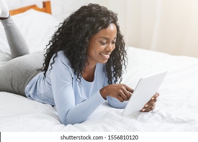 African pretty girl reading blog on digital tablet lying in bed, free space
