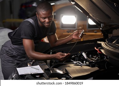 African Professional Auto Service Technician In Uniform Standing Near Car Hood Repairing And Using Check List For Car Inspect, Indoors