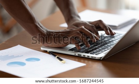 African project manager typing on pc close up view businessman hands and device, document with graphs charts lie on office desk. Business app usage, forecasting, financial report preparation concept
