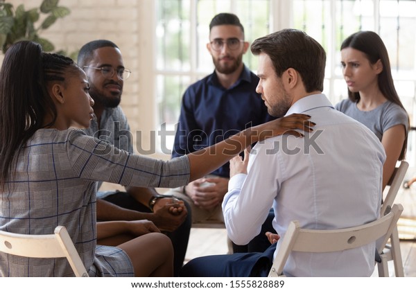 African psychologist supporting male rehab\
session participant put hand on newcomer shoulder talks encouraging\
words, medical detox center, psychological help activity struggle\
with addictions concept