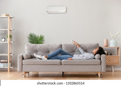 African relaxed woman in casual wear lying on sofa in living room at modern home holding air conditioner remote control, sets comfortable ideal optimal air degree temperature enjoy breathing fresh air