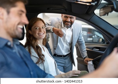 African Salesman Showing New Car To Young Couple In Showroom. Happy Man And Woman Choosing Auto In Car Dealership While Cardealer Explaining The New Technology.