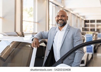 African salesman standing in car showroom near opened door of luxury car. Happy businessman buying his new suv in car salon. Smiling mature man choosing his new car. 