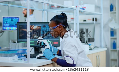 African scientist checking sample of virus using microscope in modern lab. Multiethnic team examining vaccine evolution using high tech for scientific research of treatment development against covid19