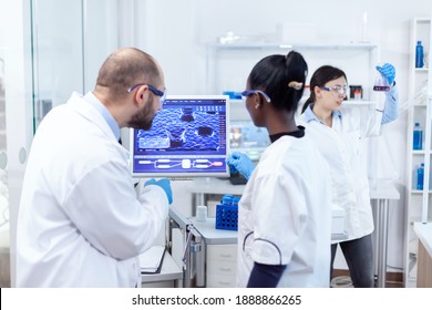 African Scientist And Doctor Studying Cure For Dangerous Virus On Computer In Busy Laboratory. Multiethnic Team Of Medical Researchers Working Together In Sterile Lab Wearing Protection Glasses And