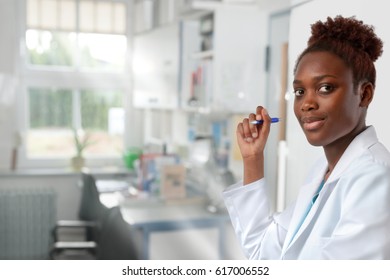 African Scientist, Medical Or Or Graduate Student. Bright, Confident Young Woman Wearing Lab Coat Holds A Pen In A Doorway Of Her Office.